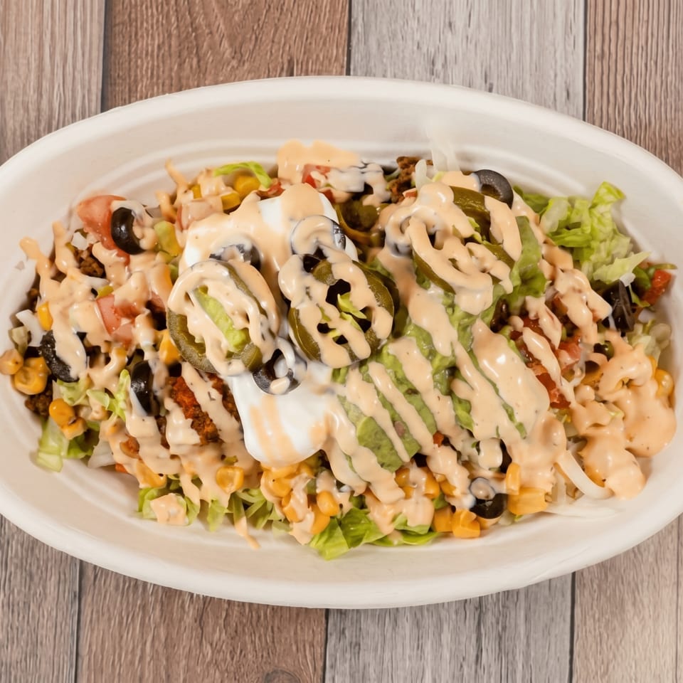 A white bowl filled with a taco salad, topped with sour cream, jalapeños, and sauce.