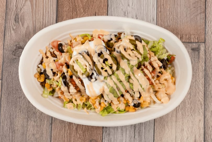 A white bowl filled with a taco salad, topped with sour cream, jalapeños, and sauce.