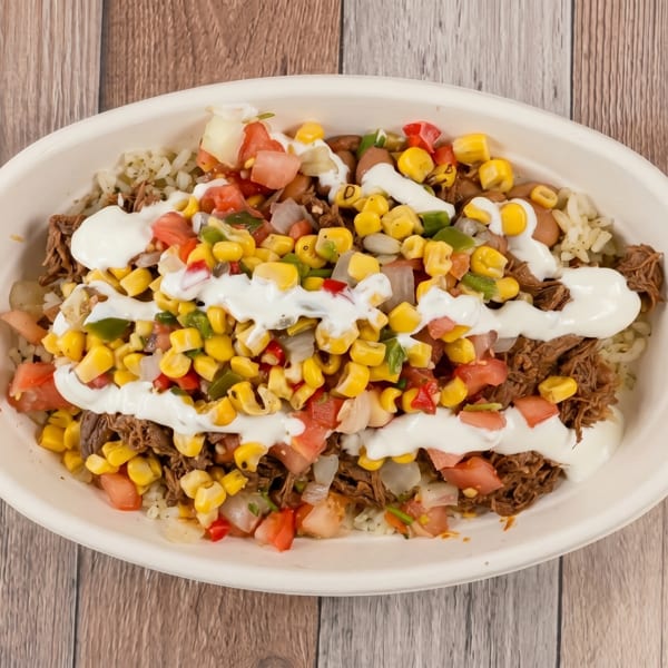 A white bowl filled with shredded meat, rice, corn, diced tomatoes, and a drizzle of white sauce.