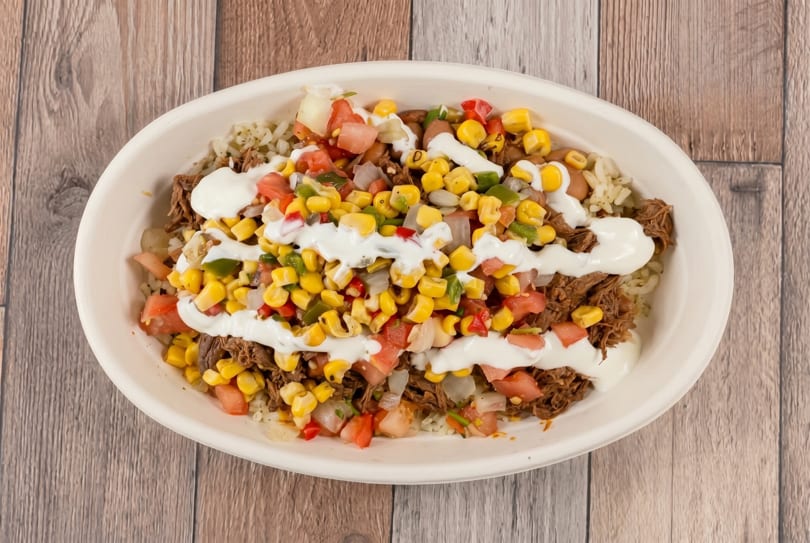 A white bowl filled with shredded meat, rice, corn, diced tomatoes, and a drizzle of white sauce.